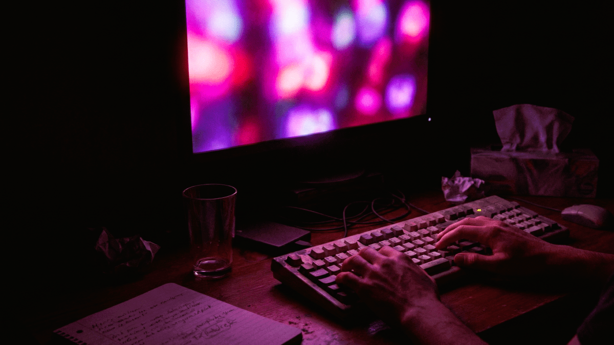 Alt Text: Dark, claustrophobic home office desk with a mechanical keyboard and glowing monitors displaying server directories. Digital noir style.